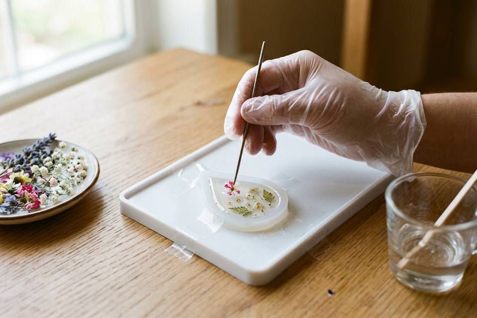 Une main gantée dépose avec une pique fine une petite fleur séchée dans la résine d'un moule en silicone en forme de goutte