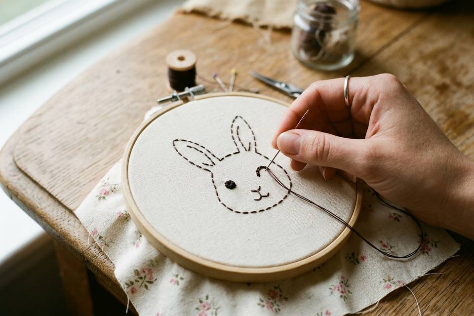 Une main brode au point arrière les yeux d'un lapin sur le tissu du devant, tendu dans un tambour